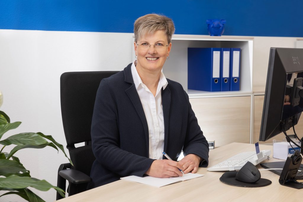 Porträtfoto einer lächelnden Immobilienberaterin in einem Büro: Sie sitzt an einem Schreibtisch, hält einen Stift über ein aufgeschlagenes Notizbuch und hat einen Computerbildschirm und Aktenordner im Hintergrund.